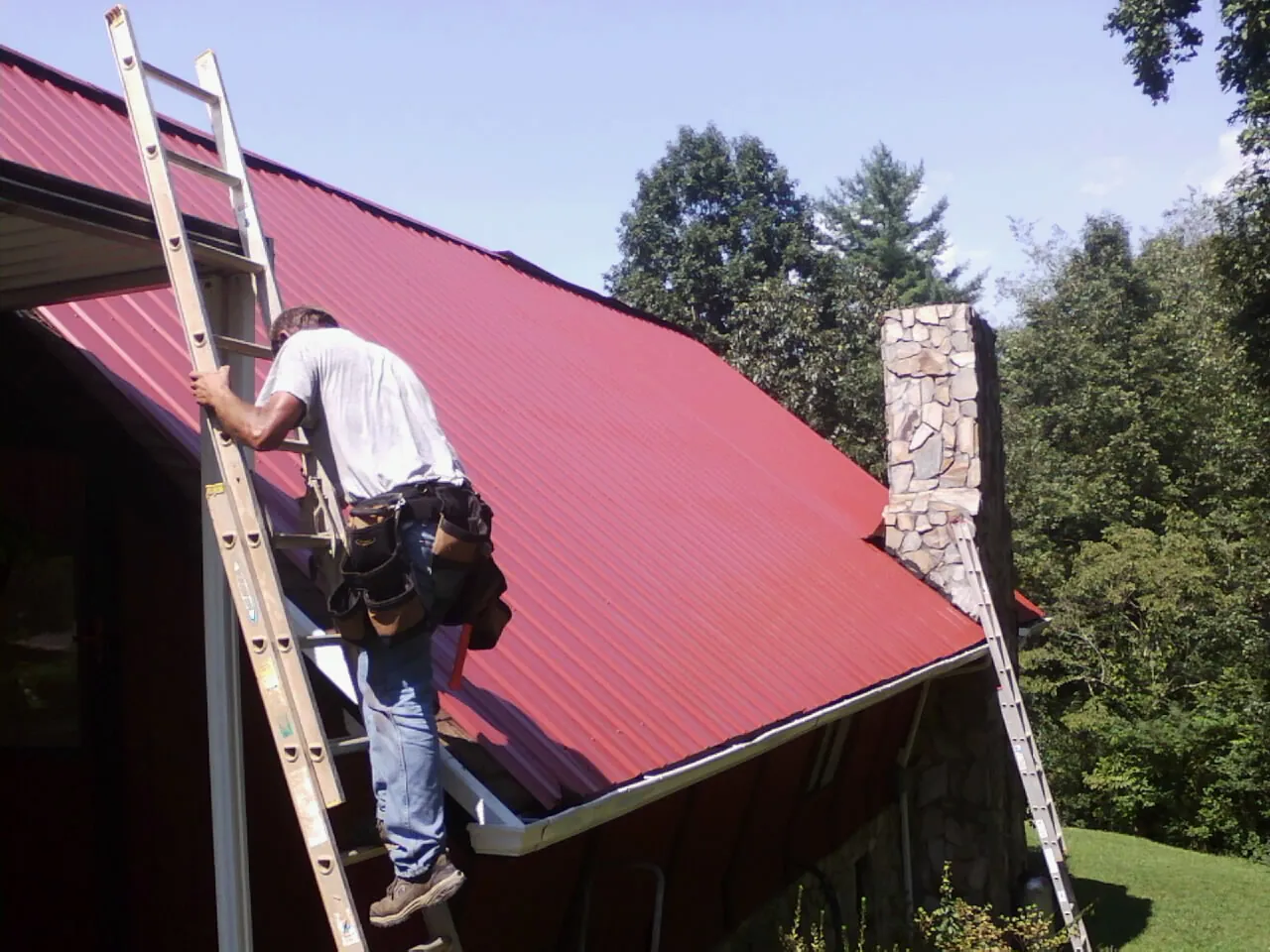 Roofer on a ladder beside a red metal roof and stone chimney — Soffit & Fascia Repair by Roofing Brothers in Harwick